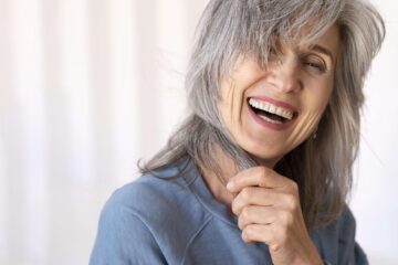 Lachende Frau Mitte 50 macht sich die Haare unter ihr Gesicht vor einem neutralen Hintergrund